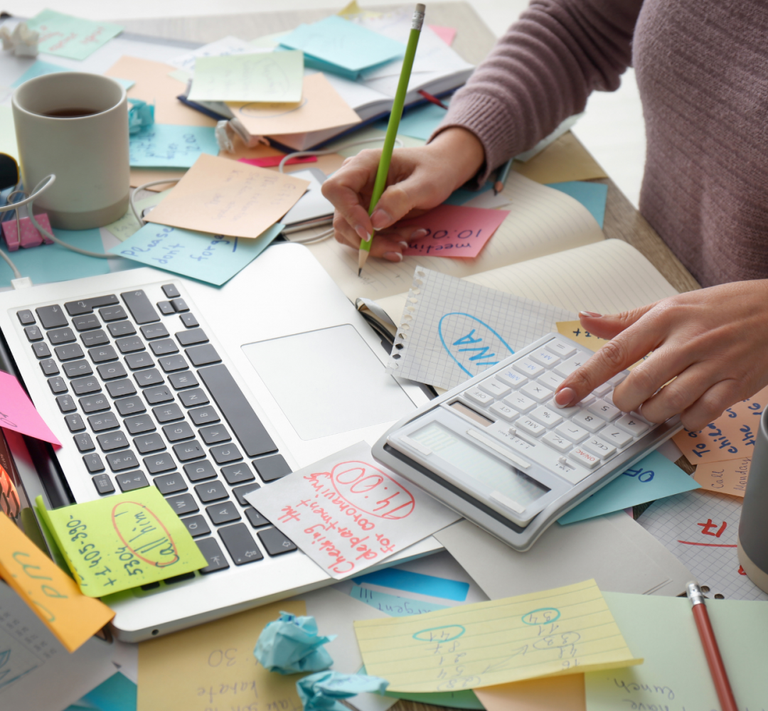 Woman working at a busy desk with laptop and notes, balancing work while retraining as a life coach