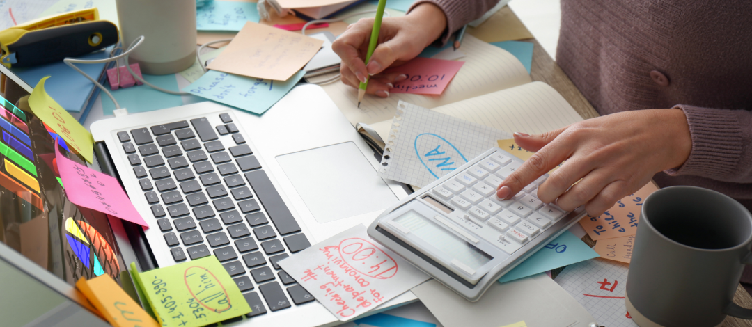 Woman working at a busy desk with laptop and notes, balancing work while retraining as a life coach