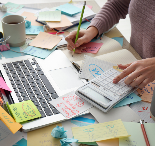 Woman working at a busy desk with laptop and notes, balancing work while retraining as a life coach