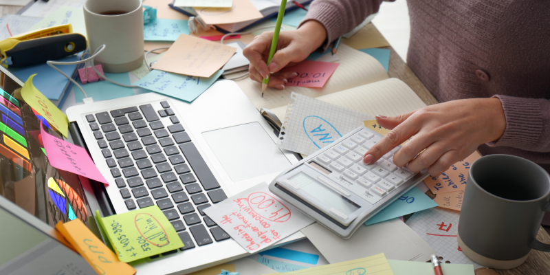 Woman working at a busy desk with laptop and notes, balancing work while retraining as a life coach